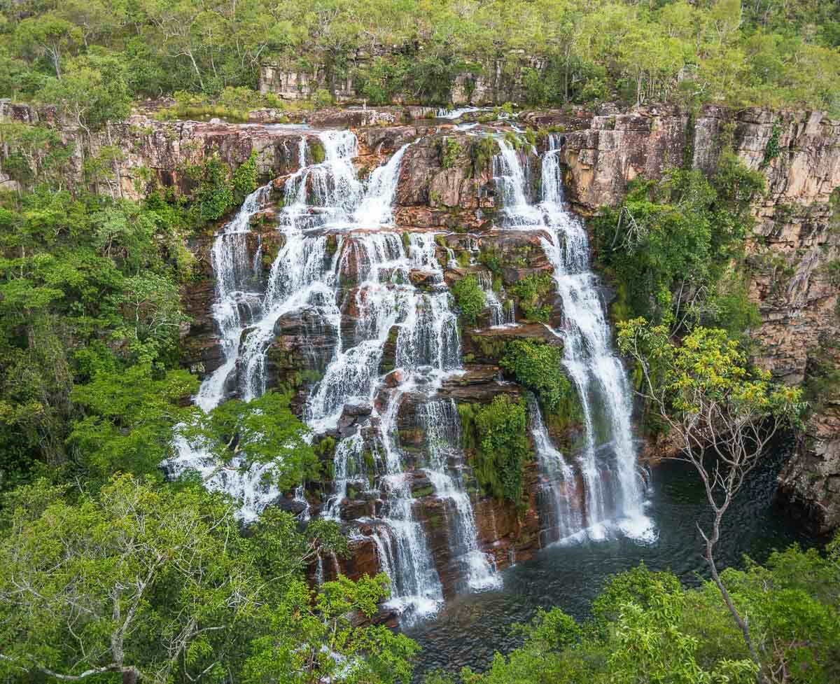 Grande cachoeira com várias quedas d’água descendo por formações rochosas na Chapada dos Veadeiros, rodeada por vegetação.