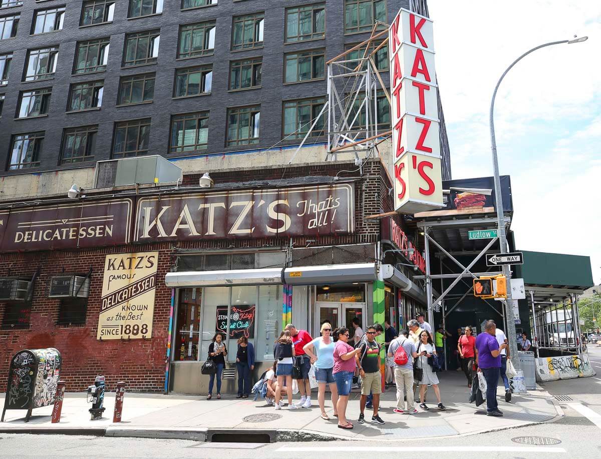 Fachada do famoso delicatessen Katz's no Lower East Side, Nova York, com uma fila de clientes na calçada. O letreiro grande vermelho e branco contrasta com o prédio de tijolos.