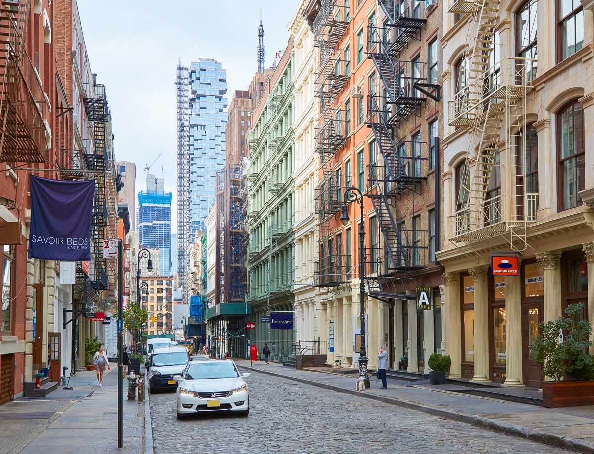 Rua do bairro Soho, em Nova York, com edifícios históricos, escadas externas de incêndio e carros estacionados ao longo da rua de paralelepípedos.