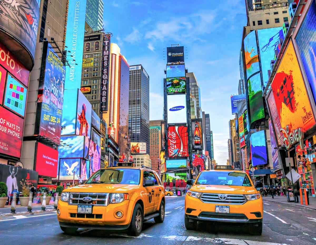 Táxis amarelos passando por uma movimentada Times Square, em Nova York, cercada por painéis publicitários iluminados e prédios altos.