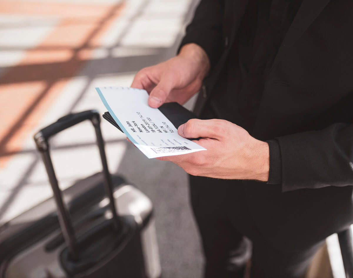Pessoa com terno preto segurando uma passagem aérea e passaporte ao lado de uma mala.