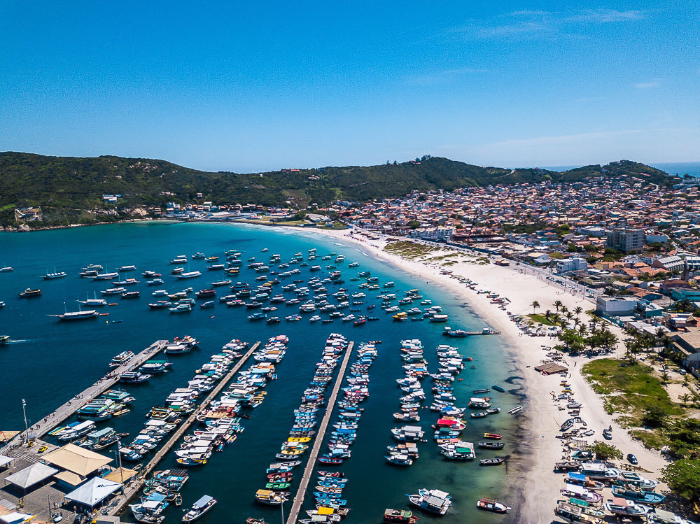 Vista aérea da Praia dos Anjos, em Arraial do Cabo, com dezenas de barcos coloridos atracados no mar azul.