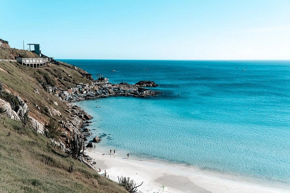 Vista da Praia Grande, em Arraial do Cabo, com mar azul-turquesa e areia branca, cercada por encostas verdes e construções no alto.