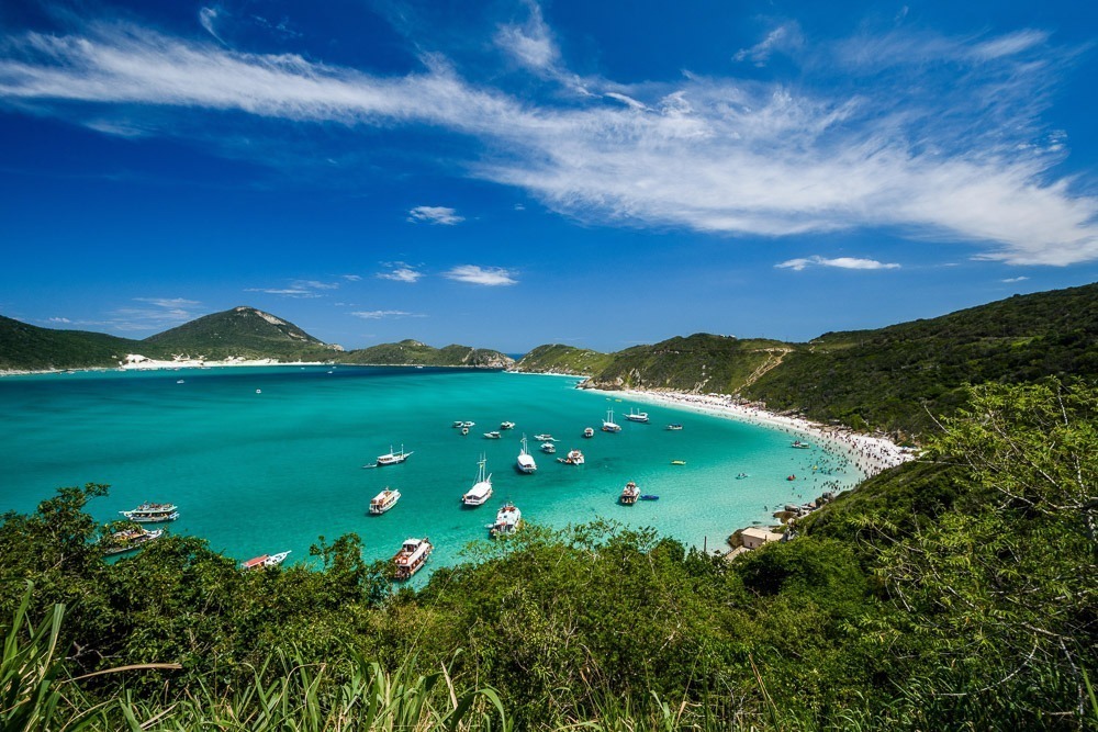 A Prainha do Pontal do Atalaia, em Arraial do Cabo, vista de um mirante, com mar azul-turquesa e barcos flutuando na água.