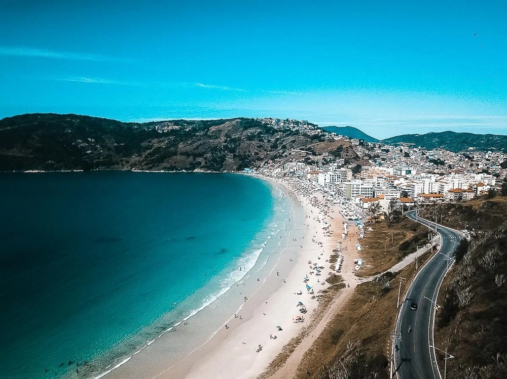 Vista aérea da Prainha, em Arraial do Cabo, com mar azul-claro e areia branca, rodeada por vegetação e edifícios da cidade.