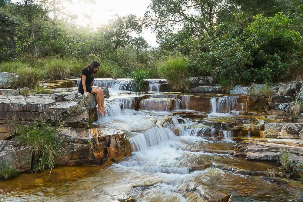 A Dri sentada em pedra na beira de uma pequena cachoeira em meio à vegetação de Capitólio.