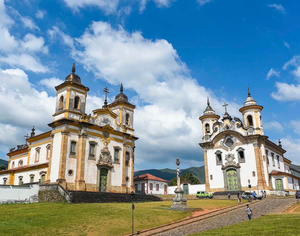 Igrejas de São Francisco de Assis e Nossa Senhora do Carmo lado a lado em praça gramada no centro histórico de Mariana, MG.