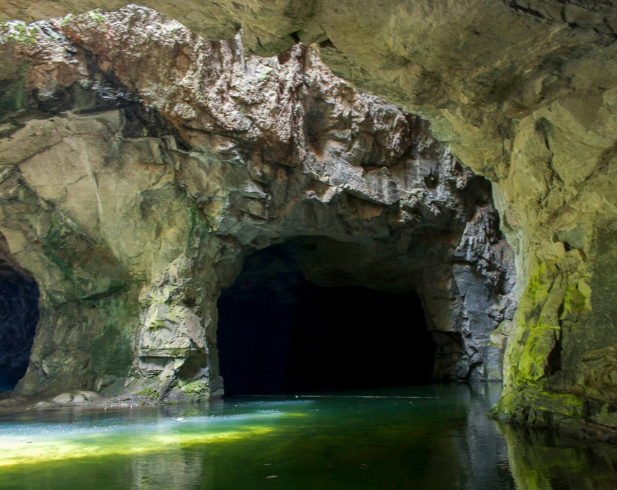 Entrada da Gruta do Anjo em Socorro, SP, com um lago de águas esverdeadas refletindo a luz dentro da caverna.