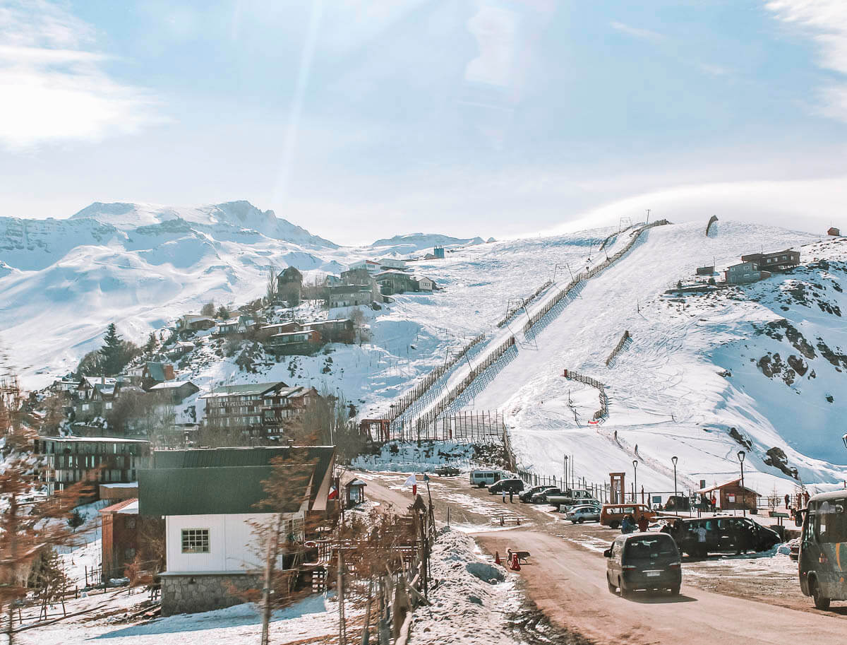 Estação de esqui Valle Nevado no Chile, com montanhas cobertas de neve, construções ao longo do morro e carros estacionados na base.