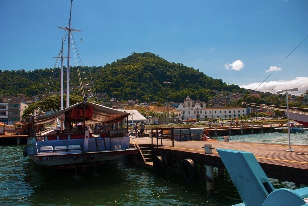 Vista do cais com barcos atracados e o centro de Angra ao fundo, em um dia de céu azul.