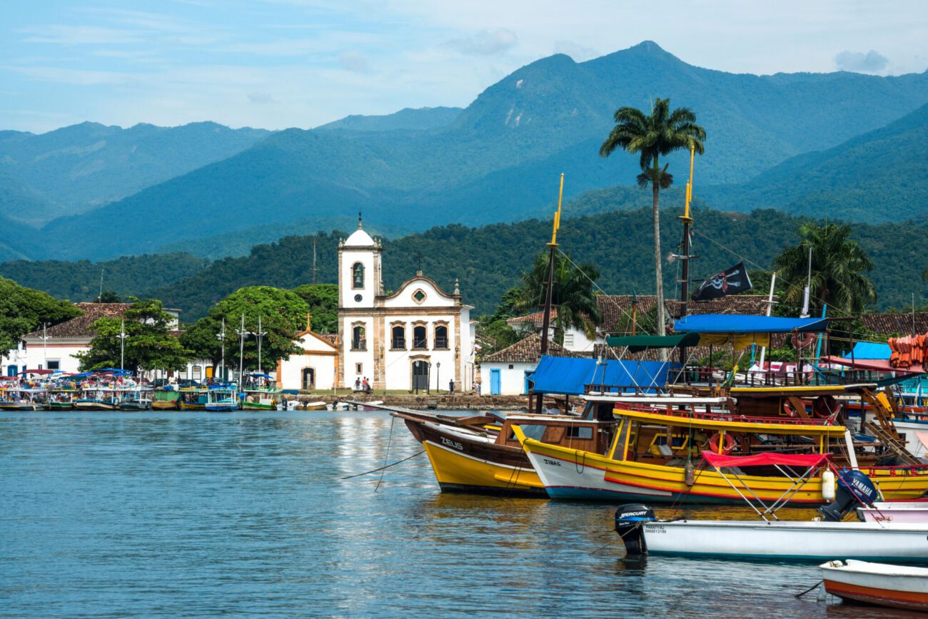 Barcos coloridos atracados em frente ao centro histórico de Paraty, com igreja colonial e montanhas ao fundo.