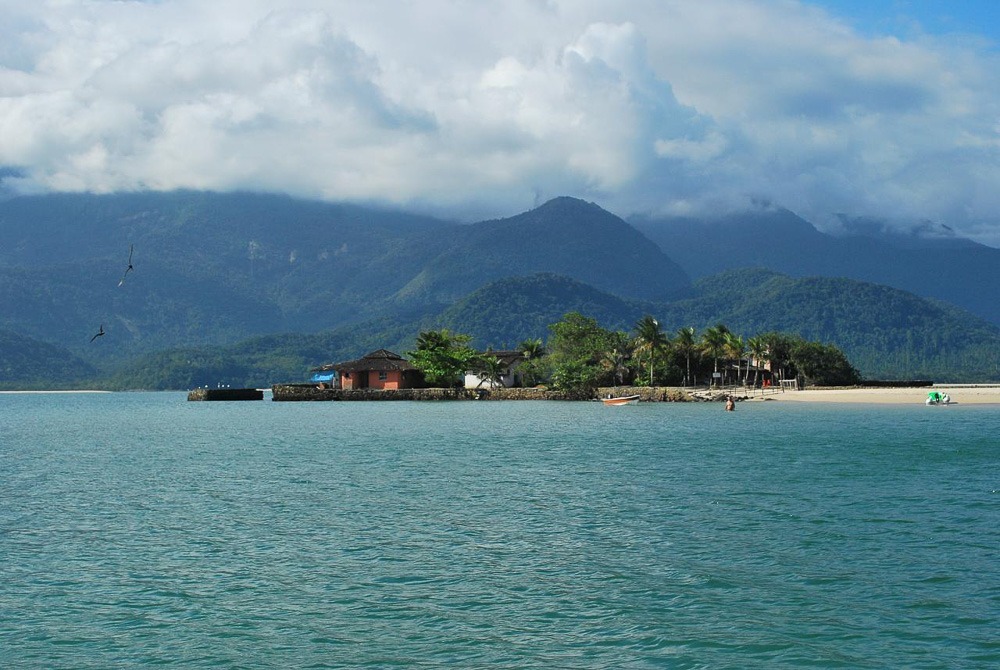 Praia com casas e árvores ao fundo, mar calmo e montanhas cobertas por nuvens.