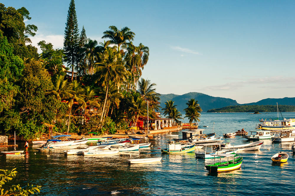 Barcos ancorados na orla da Praia do Bonfim em Angra dos Reis, cercada por muitas palmeiras e vegetação tropical.