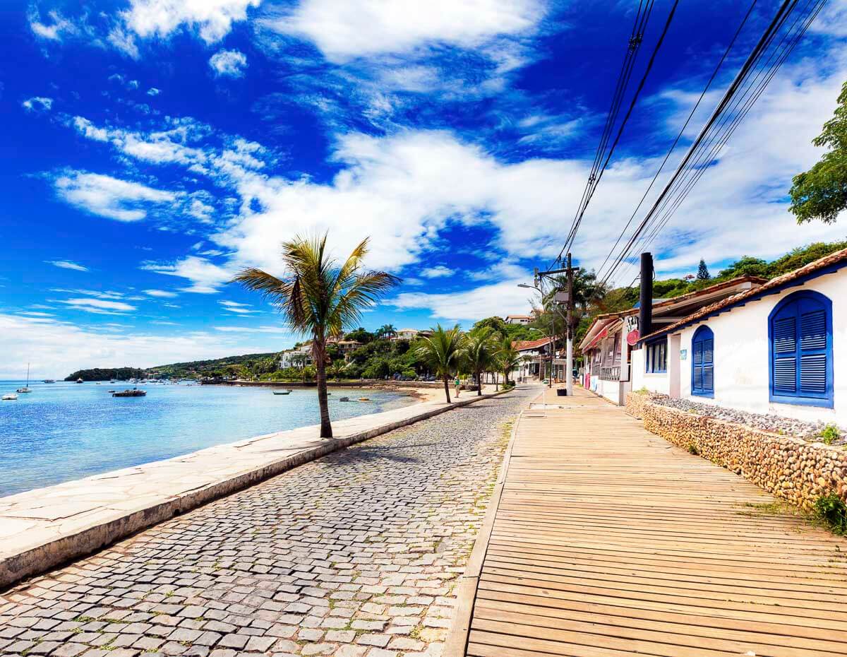 Caminho de pedras e madeira na Orla Bardot em Búzios, com casas charmosas e vista para o mar.