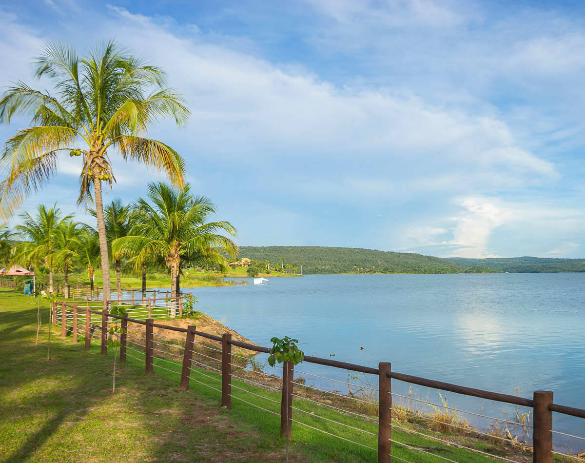 Margem do Lago Corumbá com gramado verde e coqueiros ao lado de cerca de madeira.