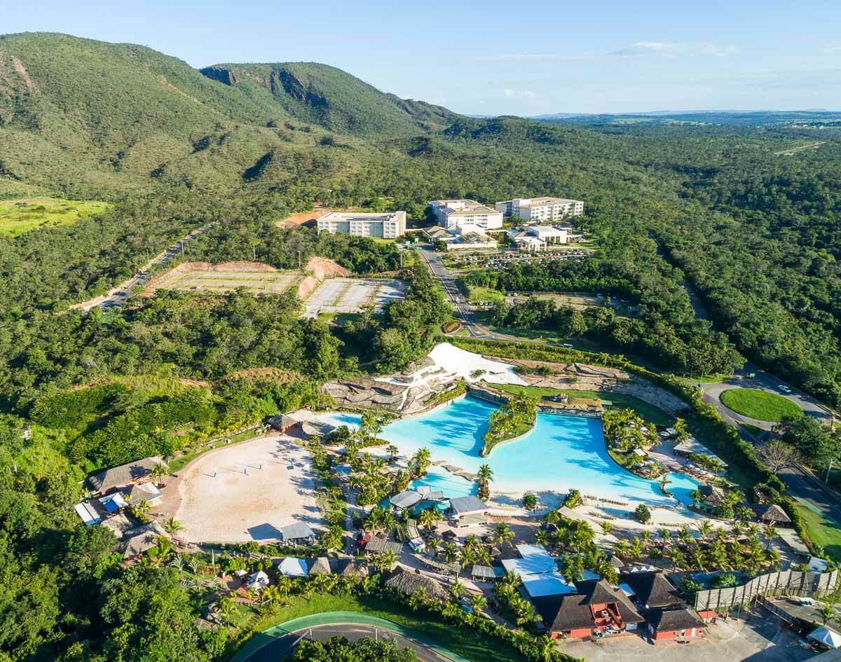 Vista aérea do complexo Rio Quente Resorts cercado por vegetação e montanhas, com grandes piscinas e área de areia.
