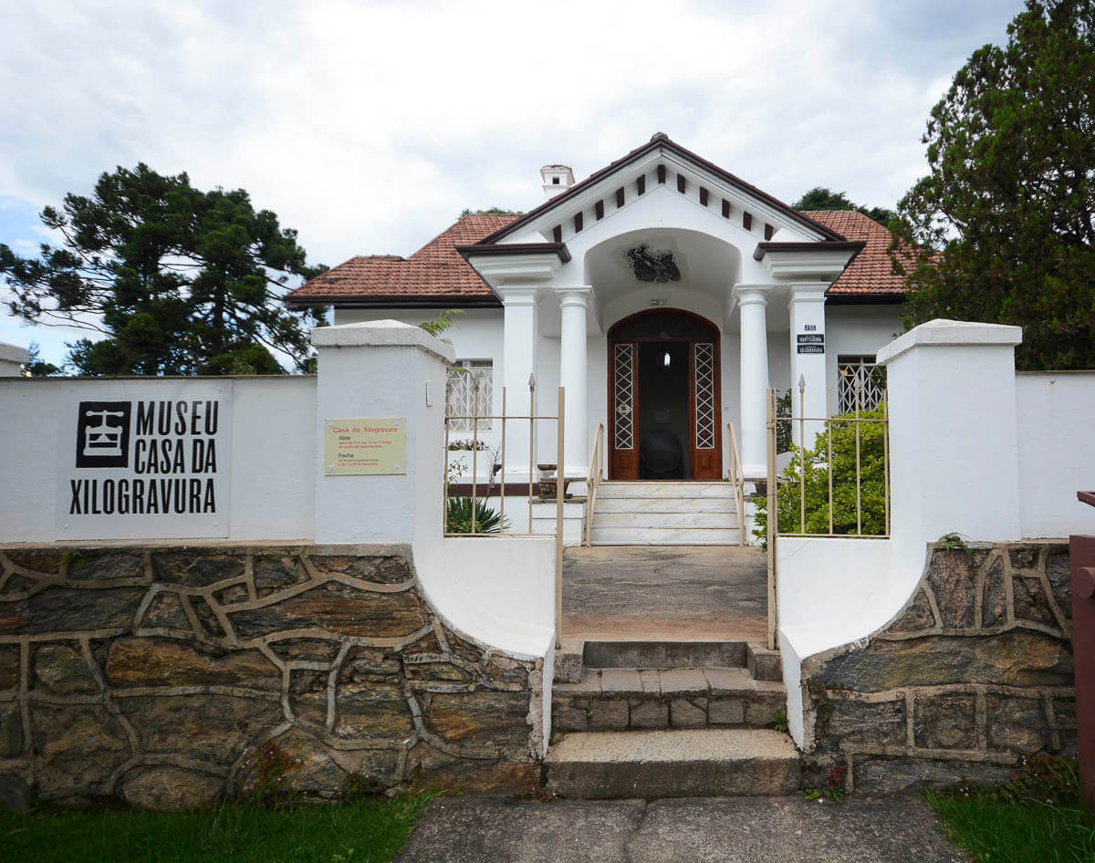 Fachada branca do Museu Casa da Xilogravura em Campos do Jordão, com portão e jardim frontal.