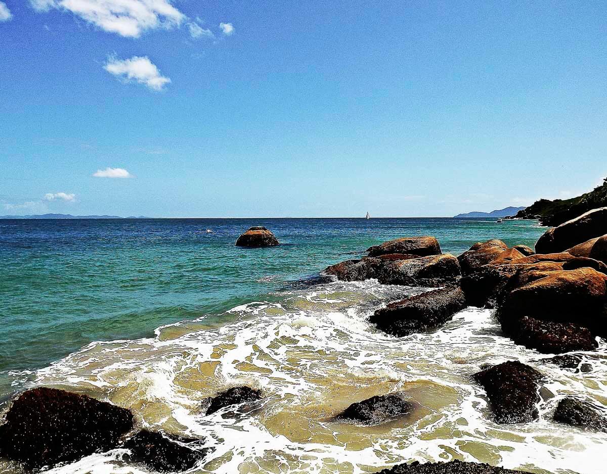 Vista da praia de Ponta das Canas com ondas quebrando entre pedras na areia.