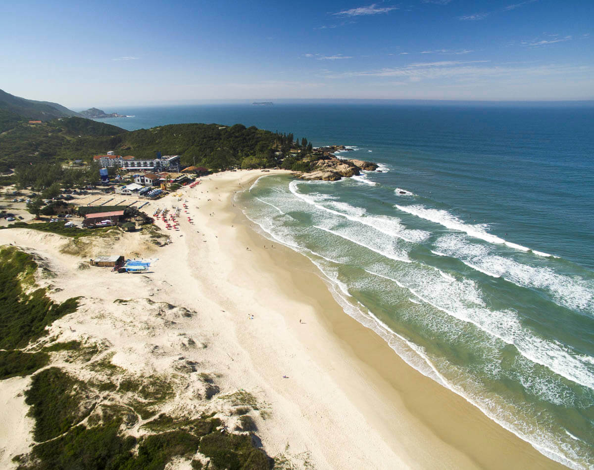Praia da Joaquina com mar azul intenso, faixa de areia clara e pedras no canto inferior direito.