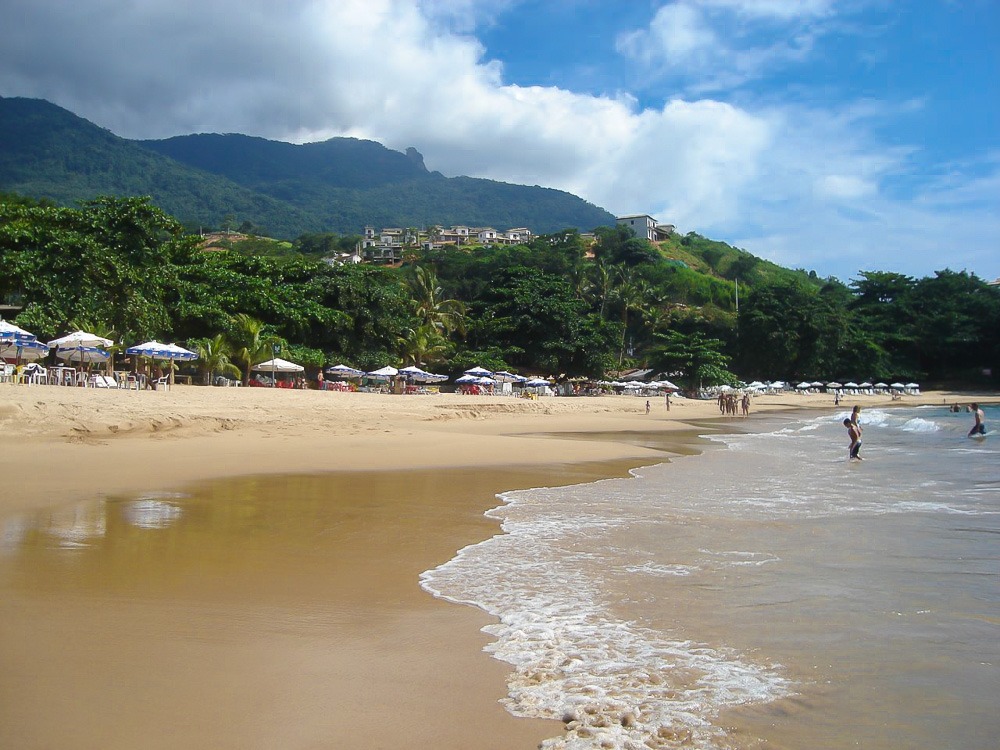 Praia do Curral em Ilhabela com cadeiras, guarda-sóis e mar calmo sob céu parcialmente nublado.