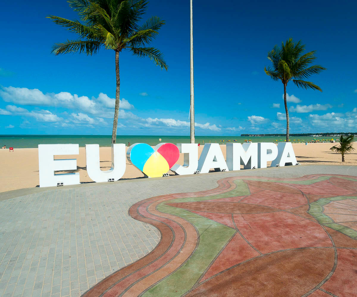 Letreiro “EU AMO JAMPA” na orla de João Pessoa, com palmeiras, calçadão colorido e mar ao fundo.