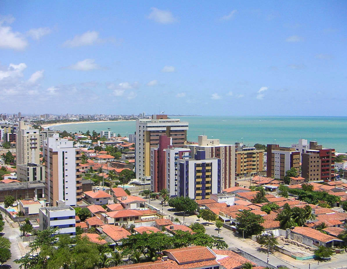 Vista da praia de Manaíra com calçadão arborizado, ciclovia, carros estacionados e mar ao fundo.
