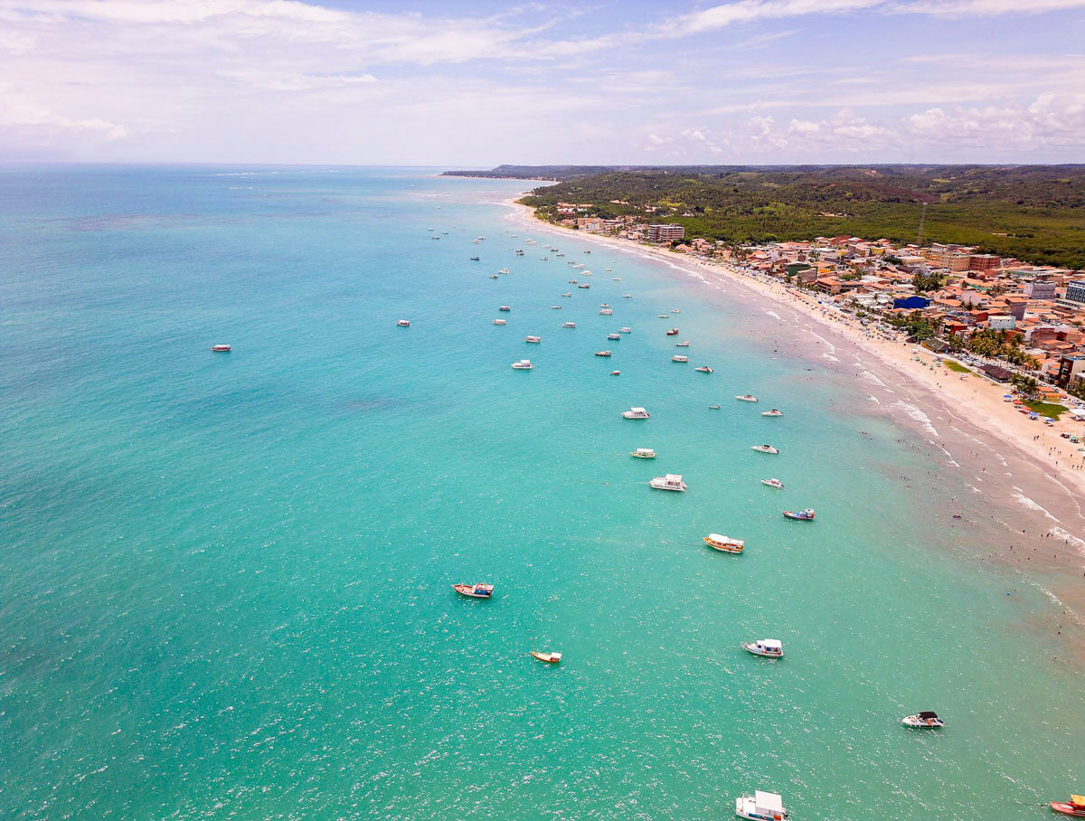 Vista aérea da orla de Maragogi com mar turquesa, dezenas de barcos ancorados e faixa urbana com prédios e casas ao longo da praia.