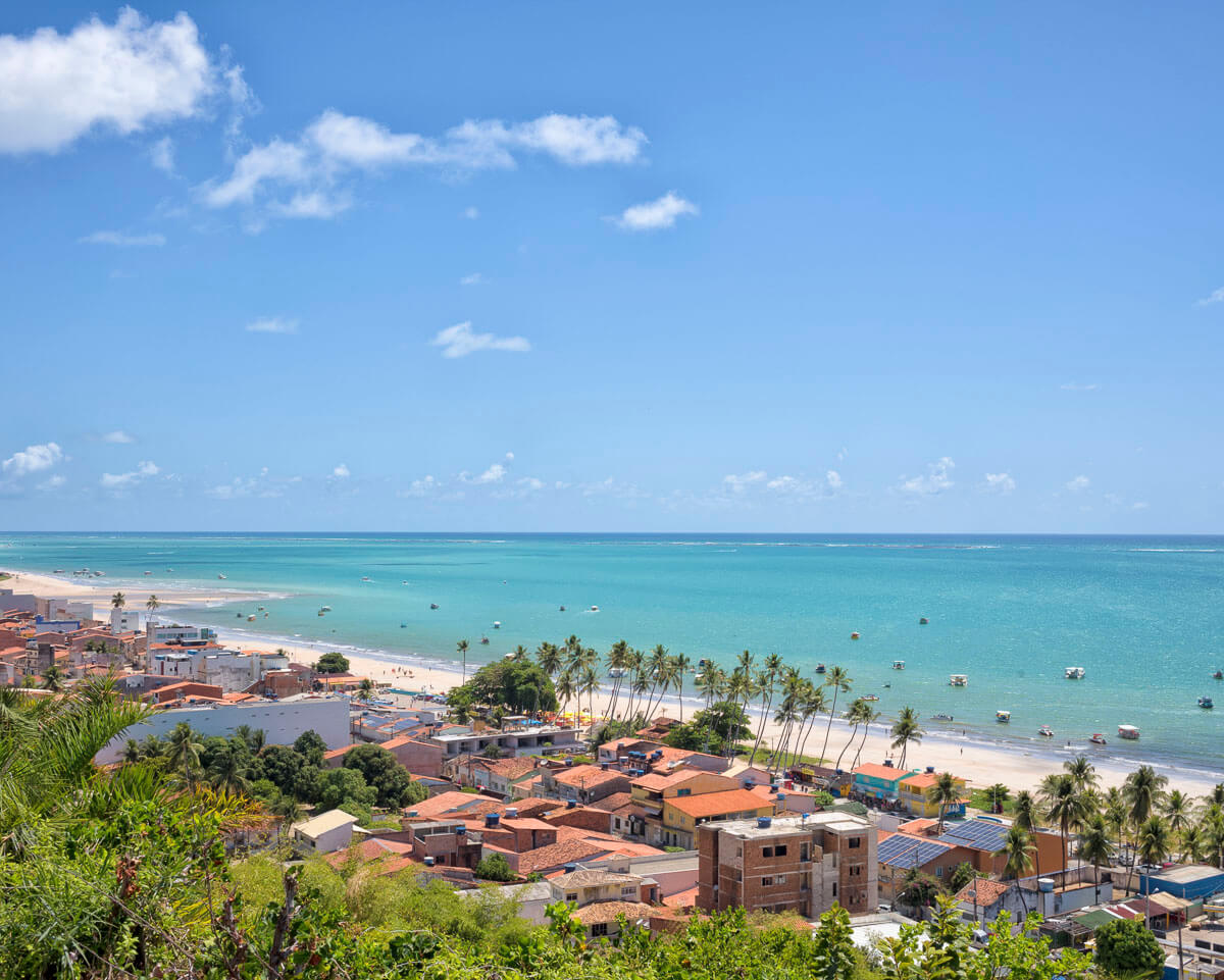 Vista ampla do litoral norte de Maragogi com mar azul-turquesa, faixa de areia clara, casas com telhados alaranjados e fileiras de coqueiros à beira-mar.
