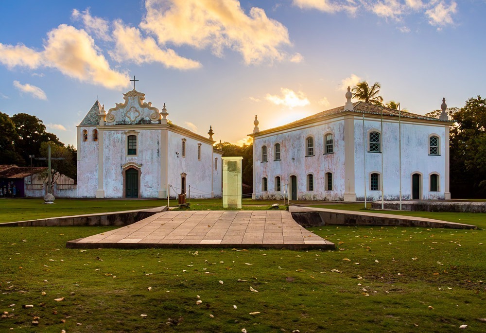 Marco do Descobrimento no centro histórico de Porto Seguro, com igreja colonial e edifício antigo ao entardecer.