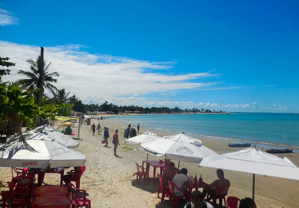 Praia de Coroa Vermelha em Porto Seguro com barracas, guarda-sóis brancos e pessoas na areia.