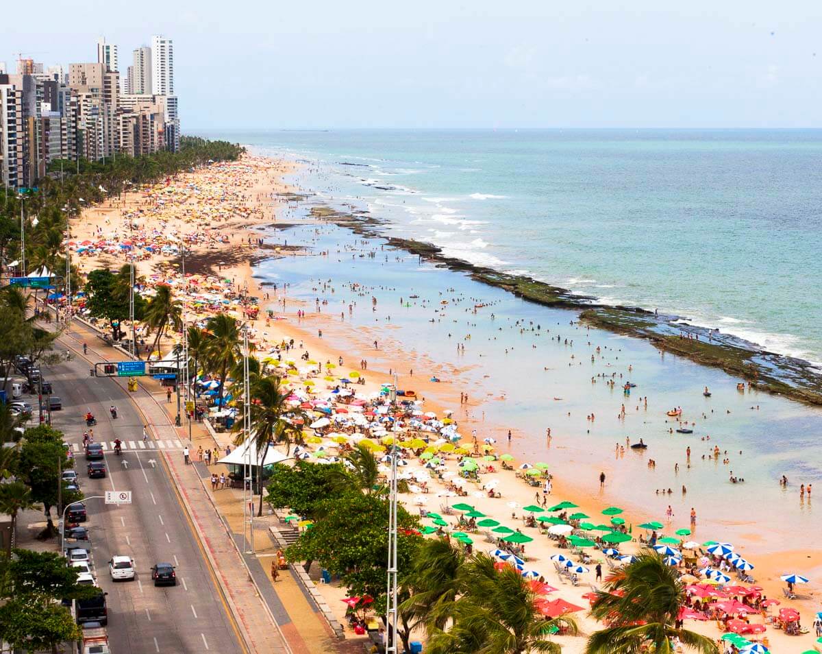 Praia de Boa Viagem lotada de banhistas, com mar azul, faixa de areia ocupada por guarda-sóis e prédios altos à beira-mar.