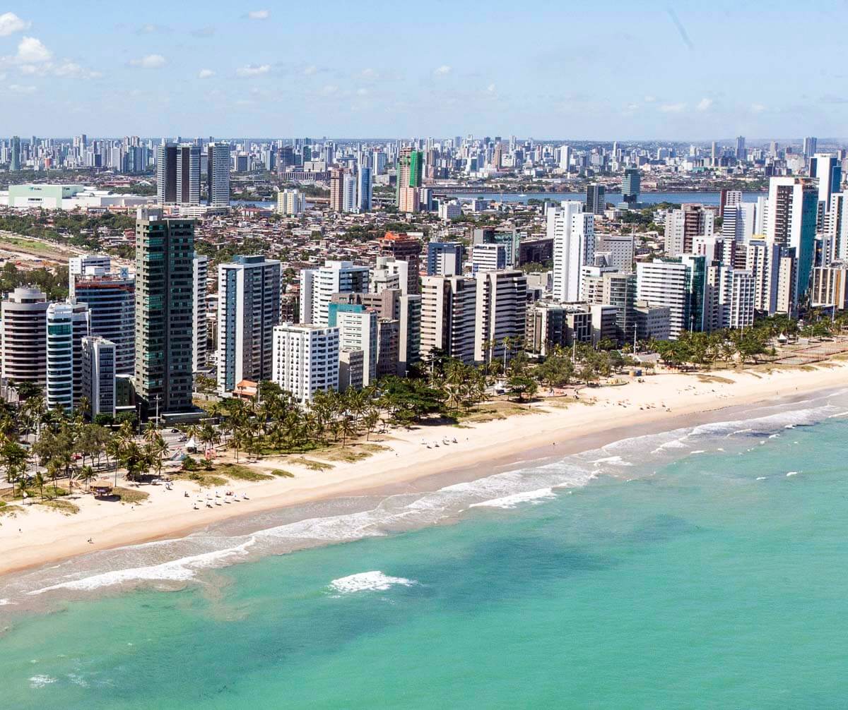 Vista aérea do bairro do Pina com praia de areia clara, mar verde e prédios altos ao longo da costa.