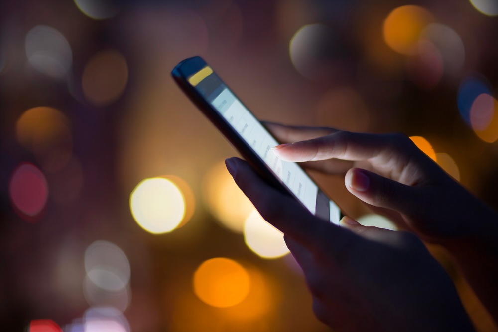 Mãos interagem com um smartphone com fundo desfocado de luzes noturnas.
