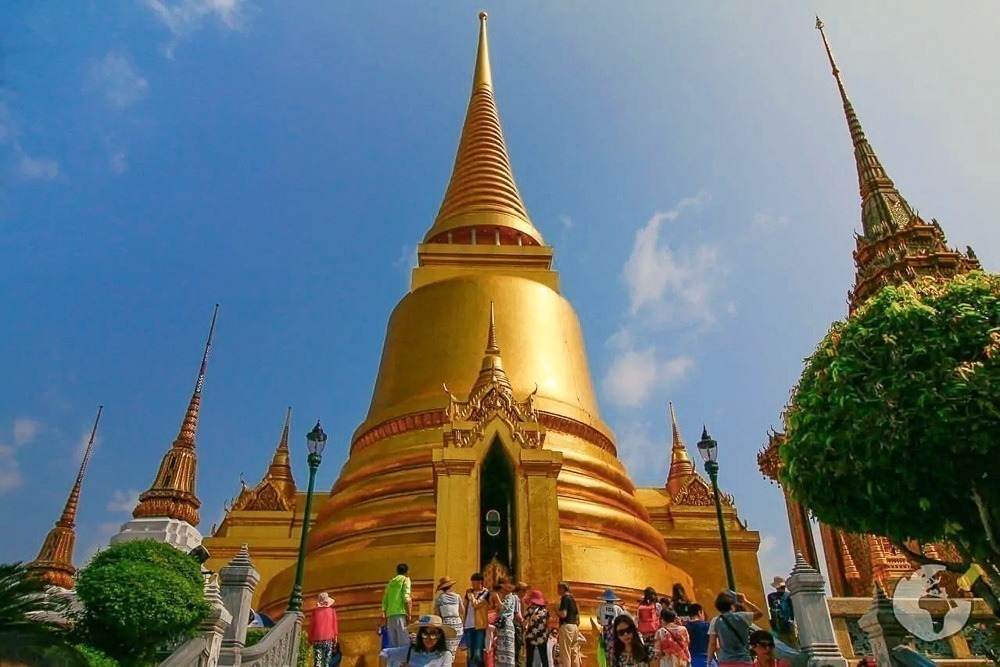 Grande estupa dourada em Wat Phra Kaew cercada por turistas e construções decoradas