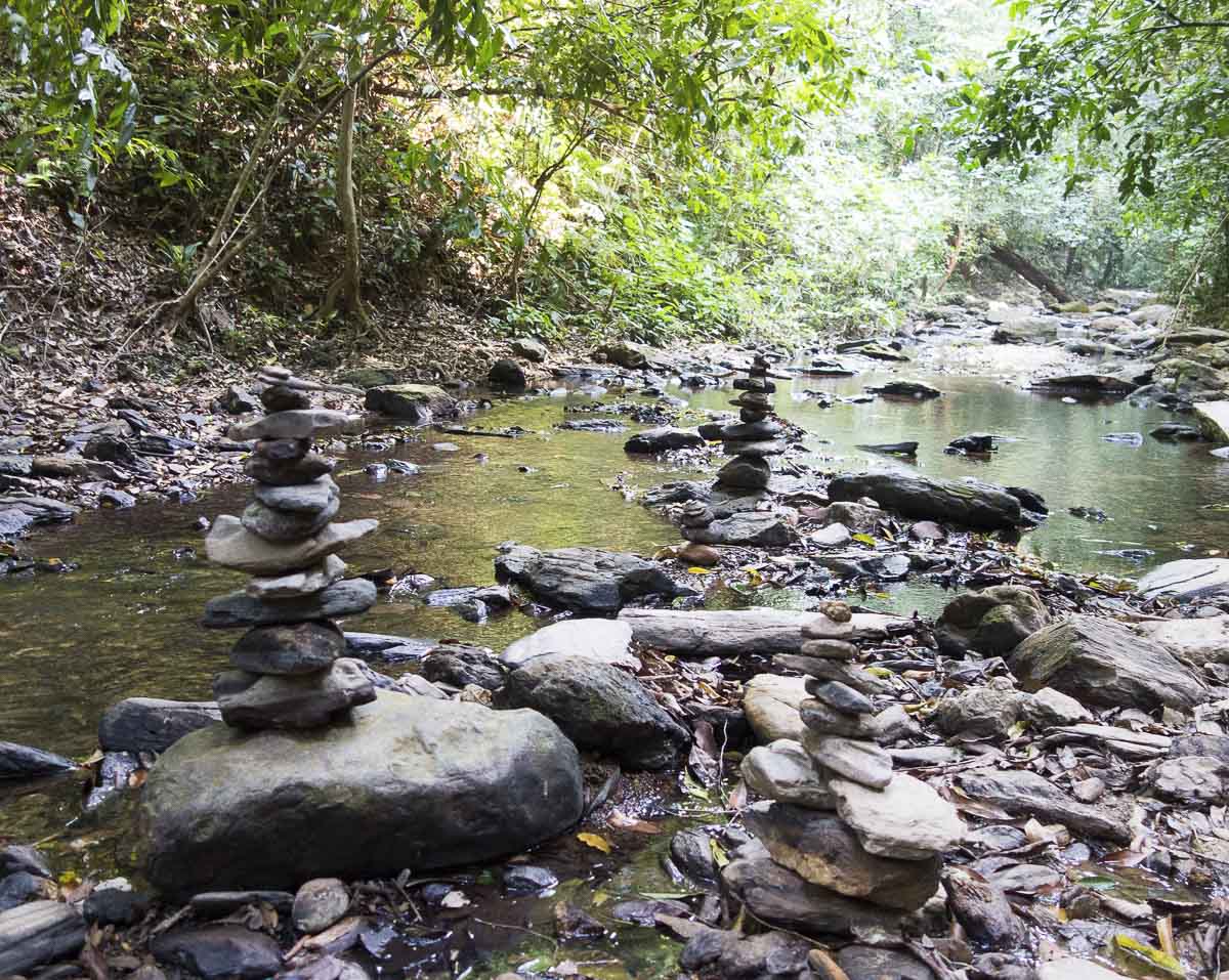 Pequeno riacho em floresta tropical com pedras empilhadas formando torres de equilíbrio.