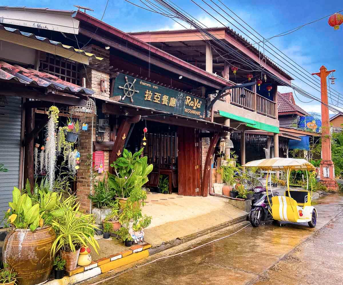 Rua com casas de madeira tradicionais, vegetação em vasos e um tuk-tuk estacionado em frente a restaurante.