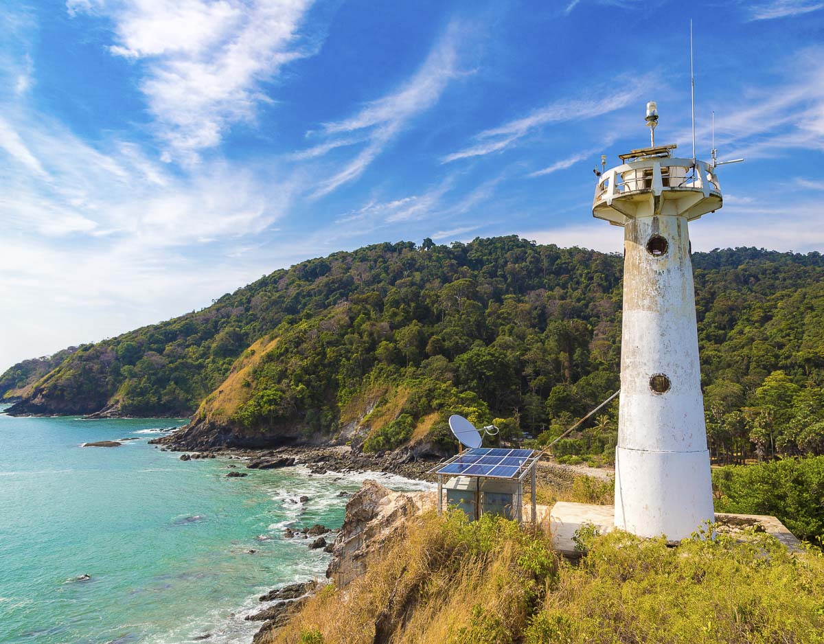 Farol branco em penhasco rodeado por floresta com vista para o mar azul-turquesa.