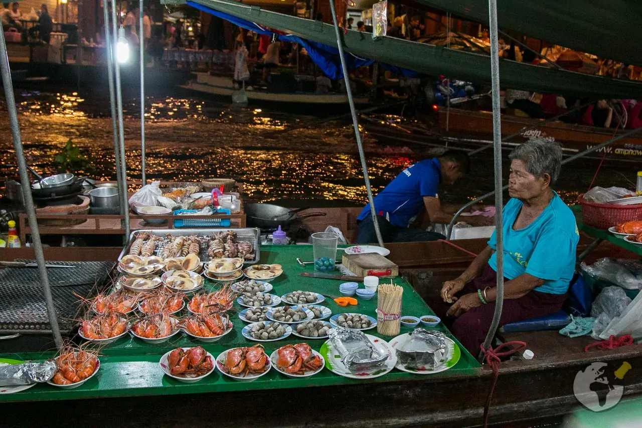 Mulher em um barco exibe pratos com camarões, mariscos e frutos do mar grelhados no mercado flutuante de Amphawa durante a noite.