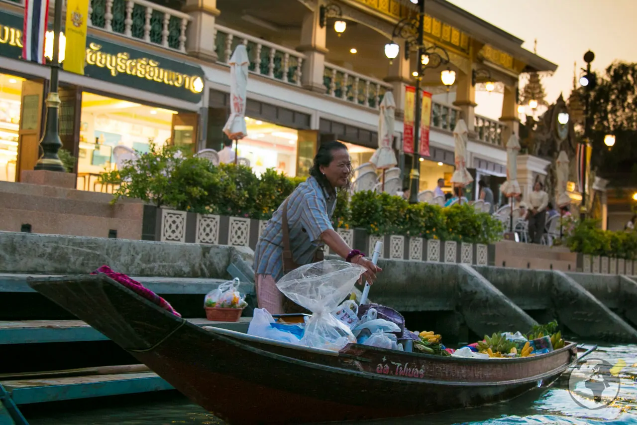 Vendedora tailandesa em um barco no mercado flutuante de Amphawa, com produtos frescos e sacolas plásticas ao redor, diante de um centro comercial iluminado ao entardecer.