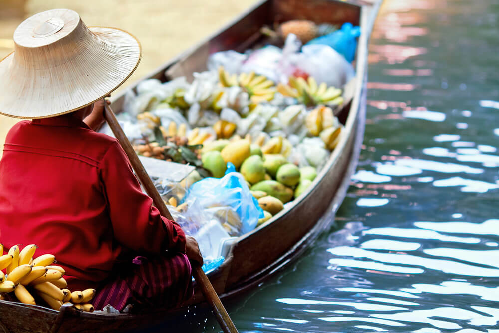 Pessoa de chapéu de palha conduz um barco repleto de frutas tropicais em canal calmo e iluminado.