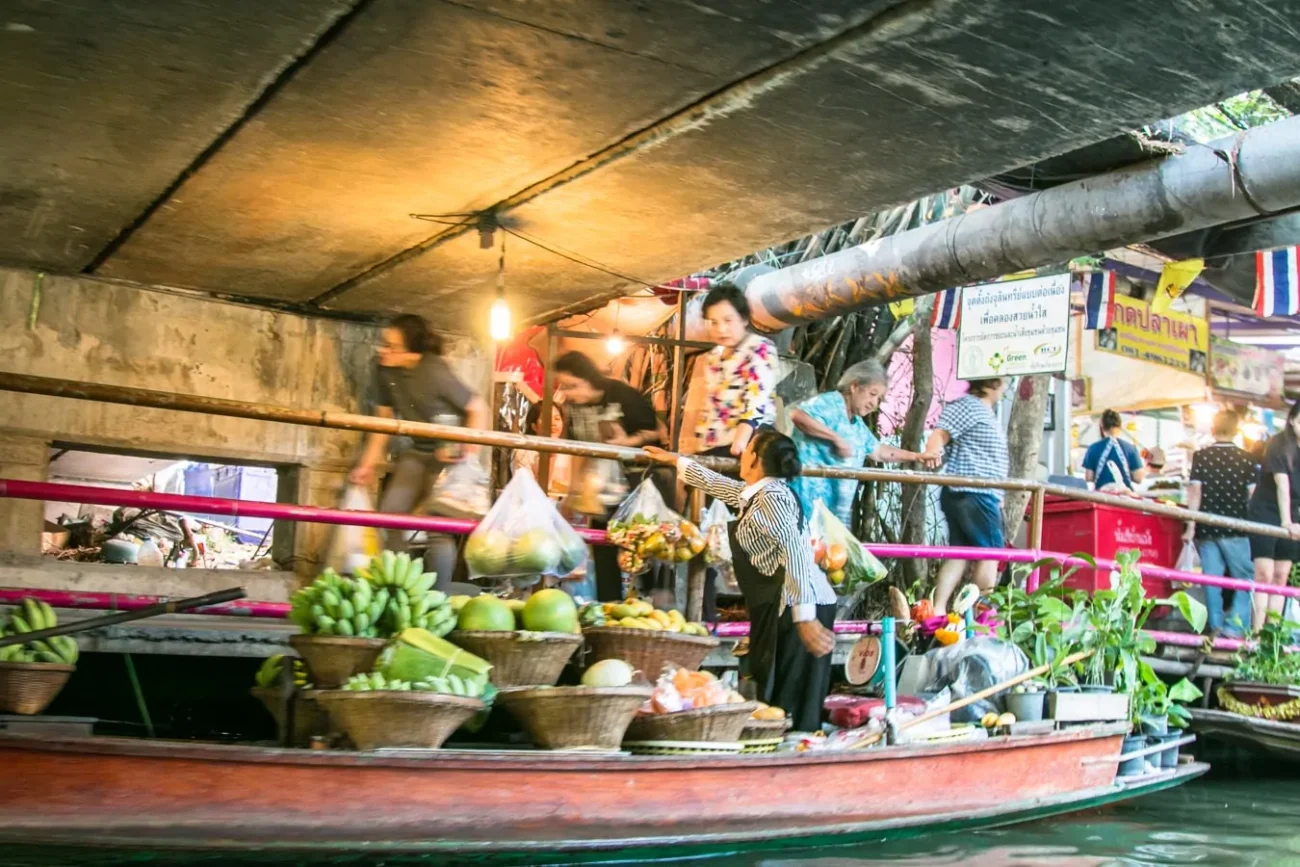 Mulher passa sacolas com frutas de barco para pessoas em calçada sob ponte de concreto.