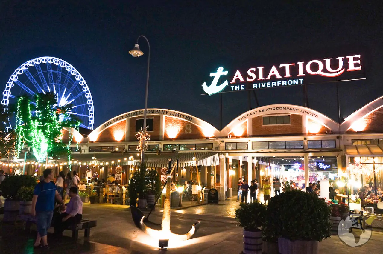 Fachada iluminada do Asiatique The Riverfront à noite, com roda-gigante ao fundo.
