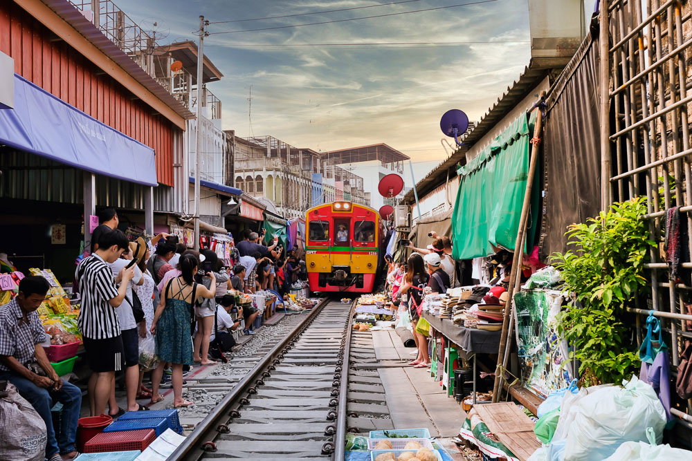Trem vermelho e amarelo avança sobre trilhos rodeados por barracas e turistas no mercado de Maeklong.