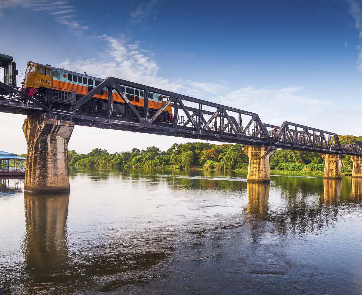 Trem passando pela Ponte sobre o Rio Kwai em Kanchanaburi com o rio e a vegetação ao fundo.