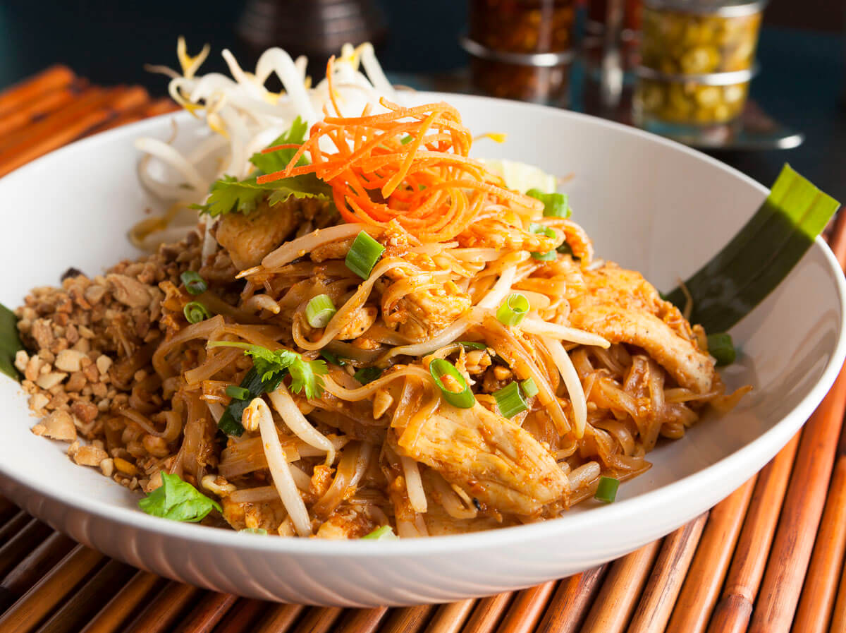 Prato de Pad Thai servido em uma tigela branca, com macarrão de arroz, broto de feijão, amendoim triturado, tiras de frango, cebolinha, cenoura ralada e coentro.