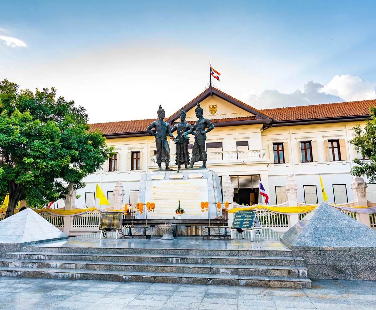 Estátuas dos Três Reis em frente a um edifício colonial com bandeira tailandesa, sede do Chiang Mai City Arts & Cultural Center.