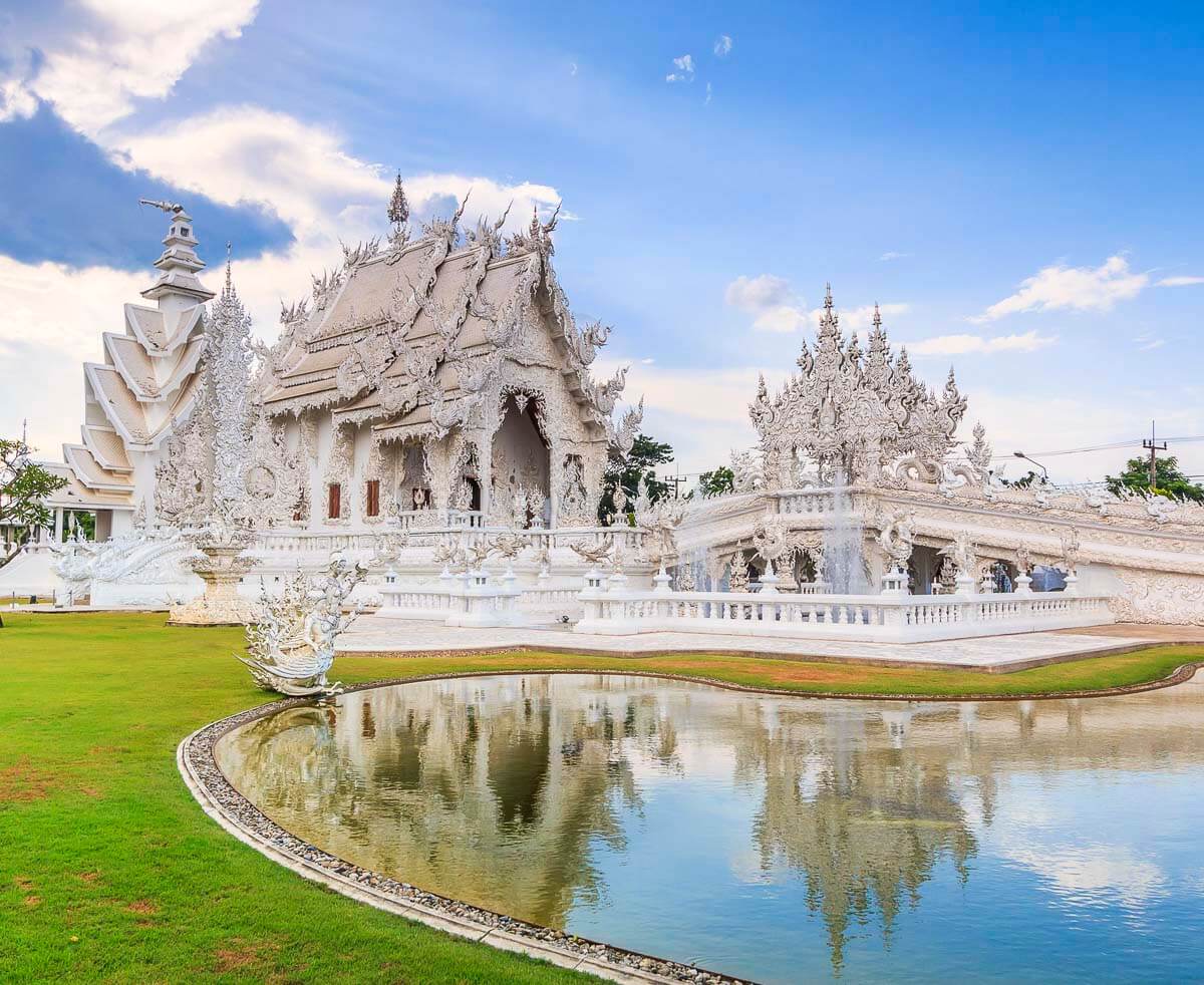 Estrutura branca ornamentada do Wat Rong Khun refletida em lago em Chiang Rai, sob céu azul.