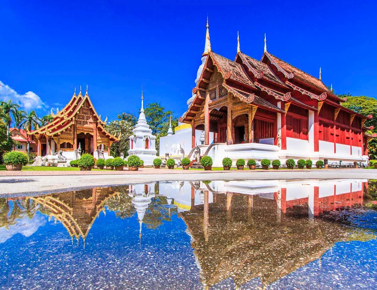Templo Wat Phra Singh em Chiang Mai com edifícios tradicionais em vermelho e dourado refletidos em uma poça d’água sob céu azul intenso.