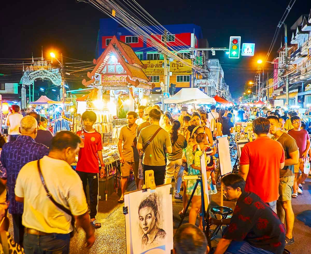 Rua movimentada com barracas iluminadas, artistas de rua e visitantes explorando o mercado noturno de sábado em Chiang Mai.