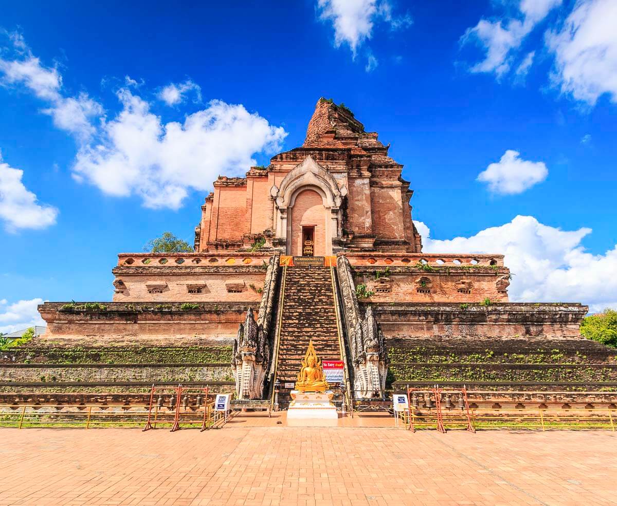 Antiga estrutura de tijolos do templo Wat Chedi Luang com escadaria central e estátua dourada de Buda ao pé.